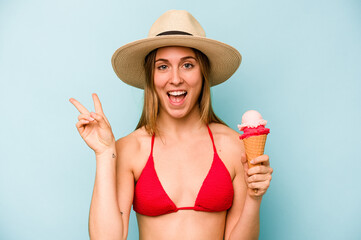 Young caucasian woman wearing a bikini and holding an ice cream isolated on blue background joyful and carefree showing a peace symbol with fingers.