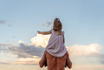 Father carrying his little daughter on shoulders with blue sky background. Happy loving family. Father and daughter child girl playing outdoors. Cute little girl and daddy. Concept of Father's day.