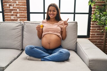 Young pregnant woman holding moisturizer oil to hydrate belly smiling happy pointing with hand and...