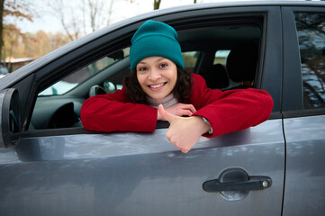Cheerful gorgeous Caucasian woman driver traveler travelling by car, leans on the driver door with opened windows and shows a thumb up, cutely smiles looking at camera.