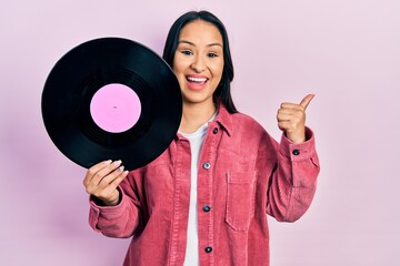 Beautiful hispanic woman with nose piercing holding vinyl disc pointing thumb up to the side smiling happy with open mouth