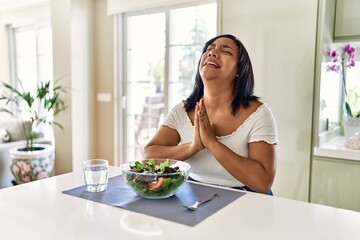 Obraz na płótnie Canvas Young hispanic woman eating healthy salad at home begging and praying with hands together with hope expression on face very emotional and worried. begging.