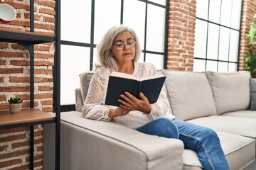 Middle age woman reading book sitting on sofa at home