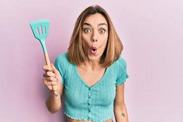 Young caucasian blonde woman holding silicone spatula scared and amazed with open mouth for surprise, disbelief face