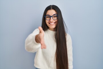 Young hispanic woman wearing casual sweater over blue background smiling friendly offering handshake as greeting and welcoming. successful business.