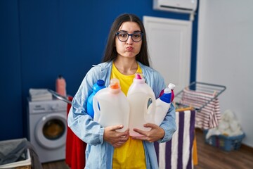 Young hispanic woman holding detergent bottles puffing cheeks with funny face. mouth inflated with air, catching air.