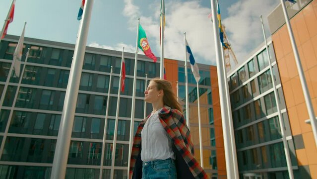Young Woman Walks and Talks by Phone in European Parliament administrative office Courtyard with Waving Flags of EU Countries in Luxembourg City. International Communications. 4K tracking shot
