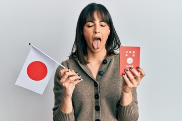 Young hispanic woman holding japan flag and passport sticking tongue out happy with funny...
