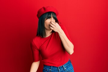 Naklejka premium Young hispanic woman wearing french look with beret smelling something stinky and disgusting, intolerable smell, holding breath with fingers on nose. bad smell