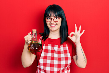 Young hispanic woman wearing cook apron holding olive oil can doing ok sign with fingers, smiling...
