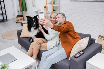 Smiling couple taking selfie on smartphone near border collie on couch.
