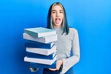 Young modern girl holding a pile of books sticking tongue out happy with funny expression.