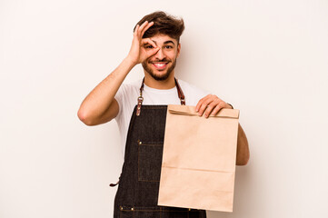 Young hispanic clerk man holding a take away bag isolated on white background excited keeping ok gesture on eye.