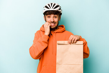 Young hispanic man wearing a helmet bike holding a take away food isolated on blue background biting fingernails, nervous and very anxious.