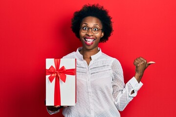 Young african american woman holding gift pointing thumb up to the side smiling happy with open mouth