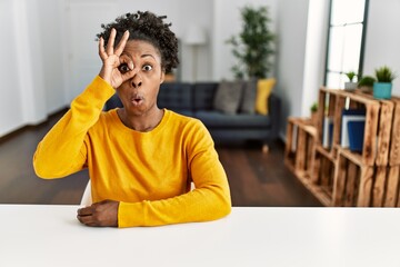 Young african american woman wearing casual clothes sitting on the table at home doing ok gesture shocked with surprised face, eye looking through fingers. unbelieving expression.