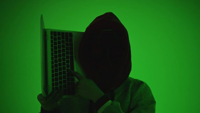 Mysterious hooded person with LED XX mask holding laptop, neon green screen background. Anonymous hacker concept