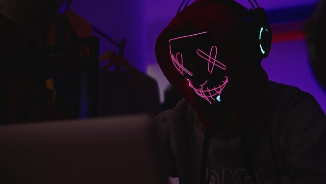 Gamer hacker in mysterious LED XX mask using computer in dark ambient room. High tech hacker gaming concept