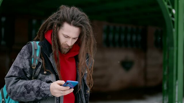 trendy man with dreadlocks is using smartphone on street in winter, swiping and scrolling display