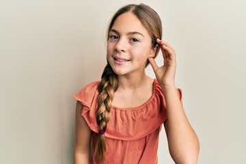 Beautiful brunette little girl wearing summer shirt smiling with hand over ear listening and hearing to rumor or gossip. deafness concept.