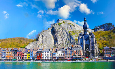 Dinant, Belgium - March 9. 2022: View over river meuse on picturesque series of old colorful houses, rock wall with citadel, gothic church against clear blue winter sky, fluffy clouds