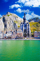 Dinant, Belgium - March 9. 2022: View over river meuse on picturesque series of old colorful houses, rock wall with citadel, gothic church against clear blue winter sky, fluffy clouds