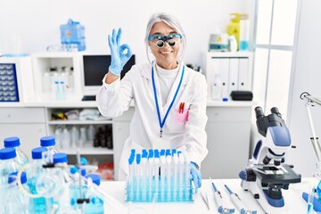 Middle age grey-haired woman wearing scientist uniform doing ok sign with fingers, smiling friendly gesturing excellent symbol