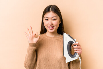 Young asian woman holding iron isolated on beige background cheerful and confident showing ok gesture.