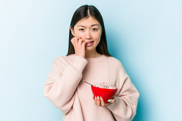 Young asian woman holding a bowl of cereals isolated on blue background biting fingernails, nervous and very anxious.