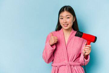 Young asian woman wearing a bathrobe and holding hairdryer isolated on pink background smiling and raising thumb up
