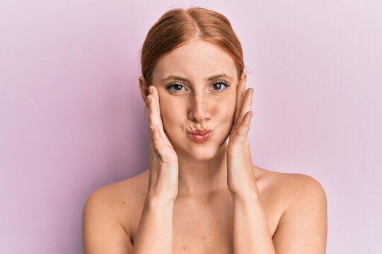 Young irish woman standing topless stretching skin face with hands puffing cheeks with funny face. mouth inflated with air, catching air.