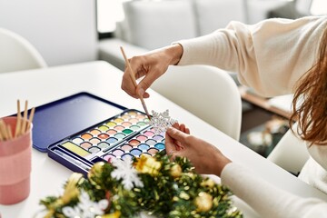Middle age hispanic woman painting star christmas decor sitting on the table at home
