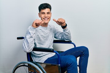 Young hispanic man sitting on wheelchair smiling cheerful showing and pointing with fingers teeth...