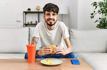 Fototapeta na wymiar Young hispanic man eating classical burger and drinking soda sitting on the sofa at home.