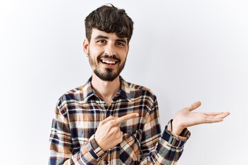 Fototapeta premium Hispanic man with beard standing over isolated background amazed and smiling to the camera while presenting with hand and pointing with finger.