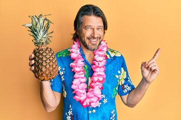 Middle age handsome man wearing hawaiian lei holding pineapple smiling happy pointing with hand and finger to the side - obrazy, fototapety, plakaty
