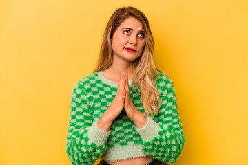 Young caucasian woman isolated on yellow background holding hands in pray near mouth, feels confident.