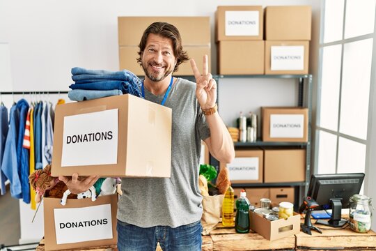 Handsome middle age man holding donations box for charity at volunteer stand smiling with happy face winking at the camera doing victory sign with fingers. number two.