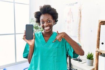 African young physiotherapist woman working at pain recovery clinic showing smartphone screen pointing finger to one self smiling happy and proud