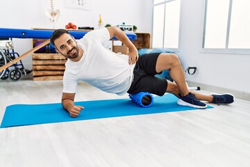 Young hispanic man patient smiling confident having leg rehab session using foam roller at clinic