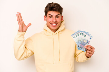 Young caucasian man holding banknotes isolated on white background receiving a pleasant surprise, excited and raising hands.