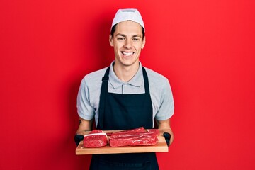 Young hispanic man holding board with raw meat smiling and laughing hard out loud because funny...