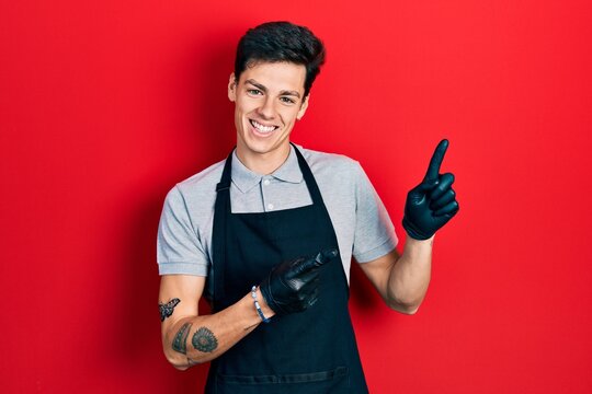 Young hispanic man wearing apron smiling and looking at the camera pointing with two hands and fingers to the side.