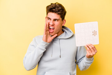 Young caucasian man holding puzzle isolated on yellow background shouting and holding palm near opened mouth.