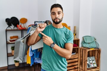 Young handsome man with beard holding shopping bags at retail shop pointing with hand finger to the side showing advertisement, serious and calm face