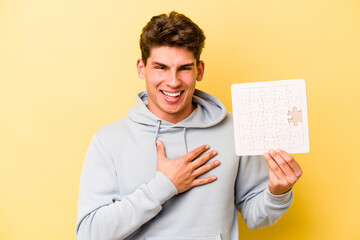 Young caucasian man holding puzzle isolated on yellow background laughs out loudly keeping hand on chest.