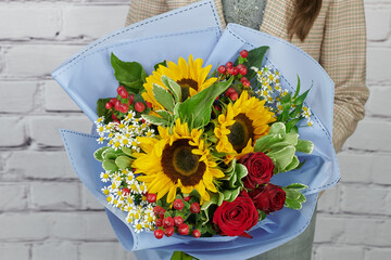 a bouquet of yellow sunflower, white daisies and red roses in a delicate blue package in the hands of a girl without a face on a light background - Powered by Adobe