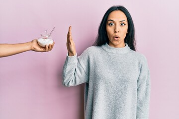 Young hispanic girl rejecting bowl with sugar scared and amazed with open mouth for surprise,...