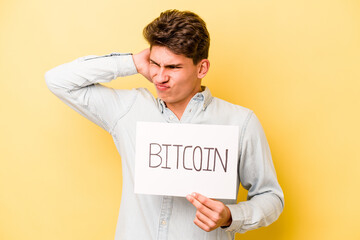 Young caucasian man holding bitcoin placard isolated on yellow background touching back of head, thinking and making a choice.