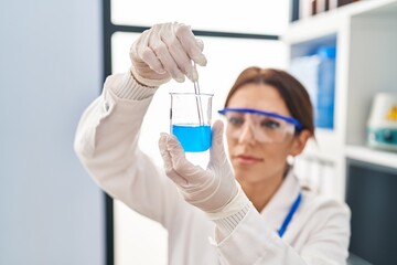 Young hispanic woman wearing scientist uniform mixing liquid on glass at laboratory